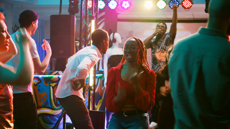 Group of people enjoying disco party on dance floor, showing funky dance moves at discotheque event. Diverse friends having fun dancing on electronic music, spotlights. Handheld shot.の写真素材