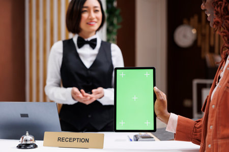 Guest using greenscreen at check in, talking to receptionist about hotel service and holding tablet. Young woman looking at isolated display with blank copyspace mockup in lobby.の写真素材