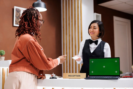 Woman giving room key card to tourist, using laptop with isolated greenscreen display on reception desk. Resort staff working at front desk with blank copyspace mockup template.の写真素材