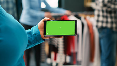 African american client looking at phone with greenscreen template, analyzing isolated display in modern boutique. Stylish woman shopping for casual wear in clothing store. Fashion conceptの写真素材