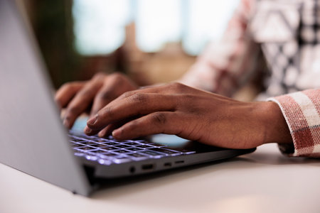Closeup of male programmer writing code on laptop keyboard while working remote from home living room. Selective focus on african american hands typing and coding software on portable computer.の写真素材