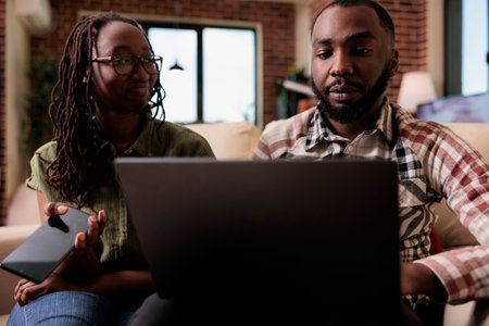 Woman holding smarthone smiling and looking at boyfriend working on laptop while sitting on couch. African american couple spending free time together using digital devices in home living room.の写真素材
