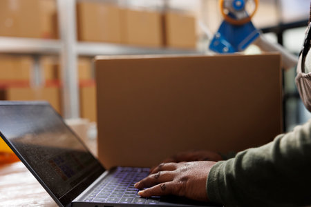 Stockroom worker checking goods quality control report on laptop computer before start working at customers order in warehouse. African american employee preparing packages for delivey. Close upの写真素材