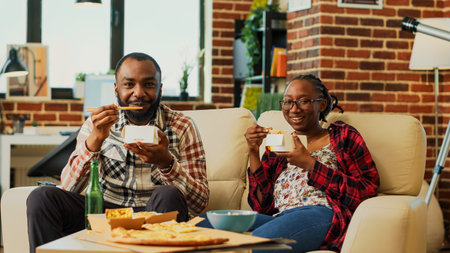 African american people eating noodles with chopsticks and binge watching action tv series at home. Young partners feeling relaxed eating asian takeaway food, watch film. Tripod shot.の写真素材