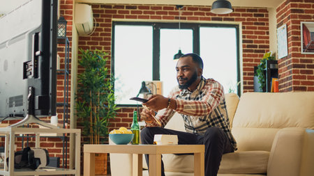 African american guy eating noodles in living room, sitting at television and watching movie. Young man serving asian food with chopsticks, ordering dinner from takeaway restaurant.の写真素材