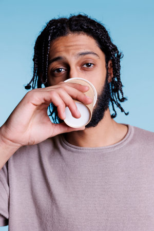 Arab man drinking coffee to go and looking at camera with calm facial expression portrait. Young person holding take out cappuccino paper cup while posing on blue backgroundの写真素材