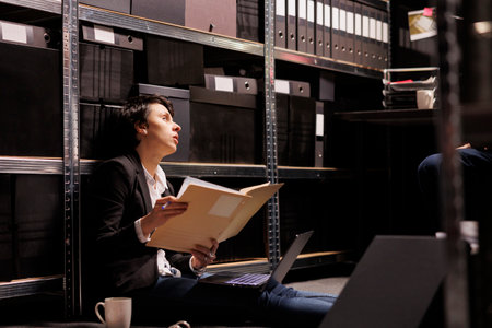 Tired private detective sitting on floor in arhive room, analyzing crime scene documents. Criminology department employees working overtime at criminal case, checking victim reportの写真素材