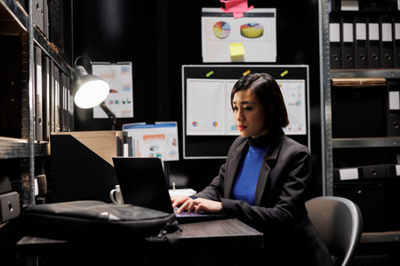 Professional businesswoman accountancy employee writing budget plan and accountancy strategy on laptop. Asian woman in file cabinet room filled with document folders and graphic chartsの写真素材