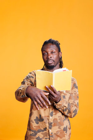 Portrait of serious reader holding literature book, reading fiction story in studio over yellow...