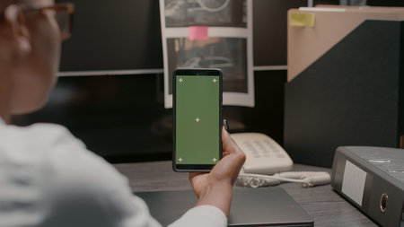 Private detective holding smartphone with greenscreen, policewoman looking at isolated copyspace display. Investigator in evidence room using chroma key mockup template in office.の写真素材