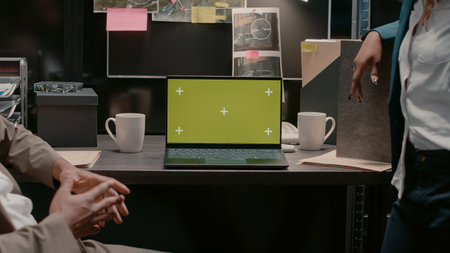 Man and woman working with greenscreen on pc, sitting in evidence room to solve crime. Team of police officers looking at laptop running chroma key template with isolated mockup screen.の写真素材