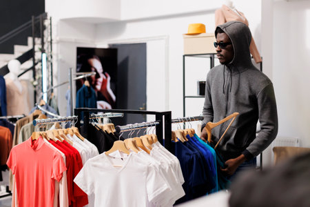 African american man thief taking a risk, looking at trendy clothes before stealing it in modern...