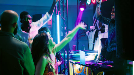Multiethnic group of adults partying and dancing on club music, having fun at discotheque with DJ on stage. Men and women jumping around dance floor and feeling happy at event. Handheld shot.の写真素材