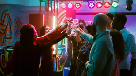 Smiling people clinking glasses filled with alcohol on dance floor, saying cheers under colorful spotlights. Young men and women enjoying cool funky party, entertainment. Handheld shot.の写真素材