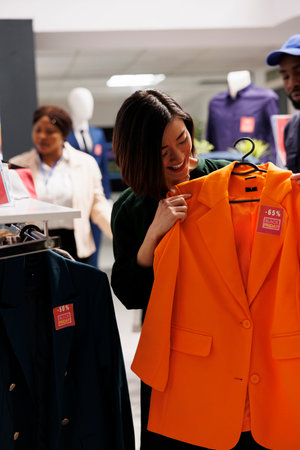 Satisfied Asian woman shopper trying on classic orange jacket with red price tag while shopping on Black Friday in clothing store. Smiling girl customer feeling happy while buying clothes on saleの写真素材