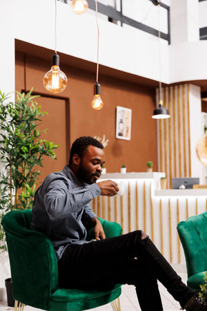 Young African American businessman sitting in hotel lobby drinking coffee, resting at lounge area waiting for business partner. Entrepreneur enjoying espresso drink during business tripの写真素材