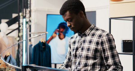 African american man shopper looking at new fashion collection, analyzing merchandise fabric in modern boutique. Shopaholic customer shopping for fashionable clothes in clothing storeの写真素材