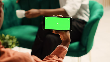 African american woman holding chroma key green screen mock up smartphone in landscape mode sitting on cozy sofa. Tourists waiting in elegant hotel lounge to be checked inの写真素材