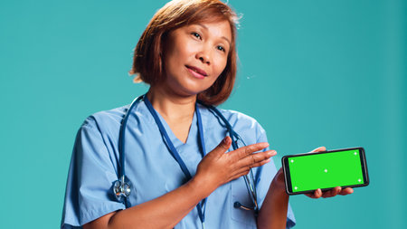 Close up shot of clinic employee isolated over studio background showing medical tape on chroma key phone green screen. Nurse holding smartphone in landscape mode, presenting informative videoの写真素材