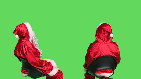 Santa claus posing on greenscreen background, young man dressed as saint nick sitting on chair and waiting. Acting impatient while he wears iconic festive costume with white beard.の写真素材