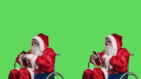 Saint nick texting and using videocall on smartphone, attend online teleconference while he is in a wheelchair dressed in red costume. Santa claus character with impairment works on phone.の写真素材