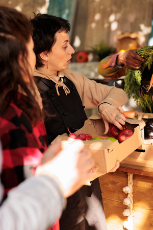 Diverse farmers selling natural fruits and veggies in boxes, serving senior man and young woman at farmers market. Smiling people looking at healthy organic colorful products.の写真素材