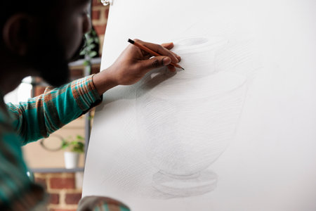 Student guy drawing with pencil on canvas. African American man artist staying focused while creating art in studio or workshop, sitting at easel sketching basic vase using pencilの写真素材