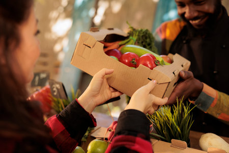 Young happy African guy farmers market vendor giving fresh organic produce box to customer,...
