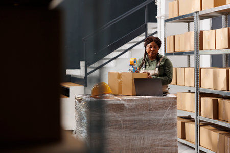 Stockroom worker preparing customers orders, using adhesive tape to pack products in cardboard boxes in warehouse. Storage room supervisor wearing industrial overall during storehouse inventoryの写真素材