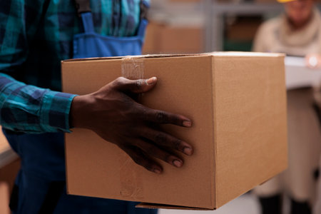 African american warehouse worker man hands holding cardboard package. Storehouse employee arms carrying carton container with adhesive tape close up, working in stockroomの写真素材
