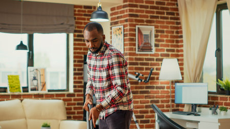 Modern male person doing spring cleaning session at home, using vacuum cleaner to clean up apartment. Young cheerful man vacuuming wooden living room floors, using washing solution.の写真素材