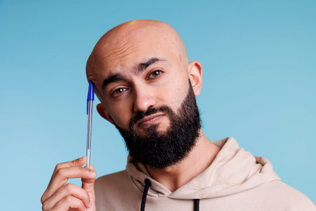 Puzzled arab man rubbing head with pen while thinking portrait. Young bald person in thought planning and solving problem while looking at camera with unsure facial expressionの写真素材