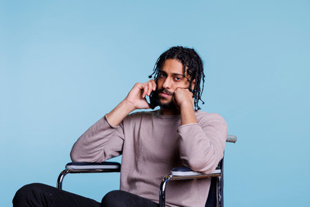 Calm arab man with disability sitting in wheelchair and talking on smartphone while looking at camera. Young person with physical imparment chatting on mobile phone portraitの写真素材