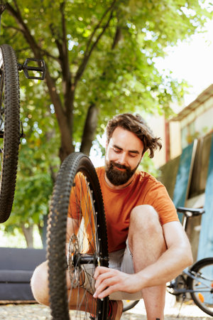 Active and committed man crouching to repair bike wheel rim in home yard using professional tool. Healthy male cyclist working on dismantled bicycle tire outside using specialized wrench.の写真素材