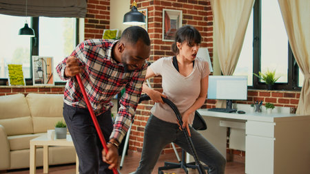 Diverse couple showing dance moves and having fun cleaning apartment, using mop to sweep dust and vacuum at home. Life partners dancing and singing, using washing solution, spring cleaning.の写真素材