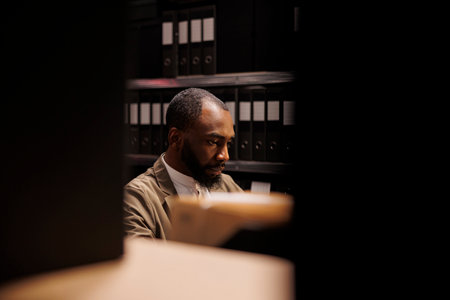 Private detective searching information in police records and working late. Policeman sitting in room with racks full of folders with case files and solving crime at night timeの写真素材