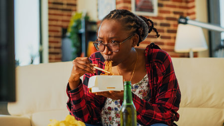 Female person using chopsticks to eat asian food, laughing at favorite film on television. Young woman eating noodles in delivery box and drinking beer, having fun at television. Handheld shot.の写真素材