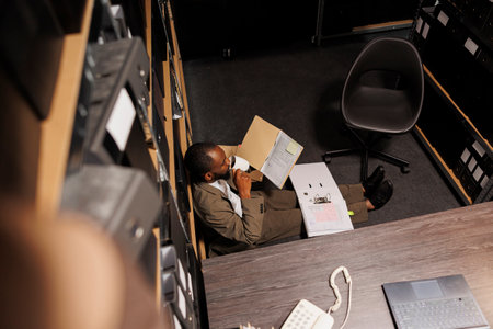 African american cop drinking coffee and analyzing police records on floor. Law enforcement professional working overtime on complex crime case and reading csi expertise reports top viewの写真素材