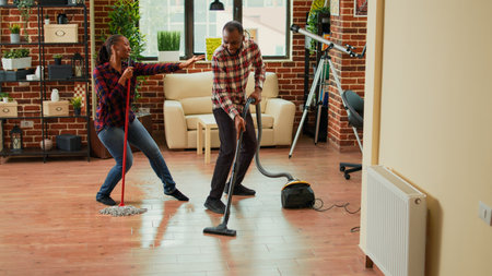 Young happy couple listening to music and washing floors in apartment, doing silly dance moves. Cheerful people doing spring cleaning and dancing, mopping tiles. Handheld shot.の写真素材