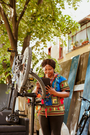 Energetic and enthusiastic black woman diligently repairing and upkeeping various bike components using digital tablet in home yard. Young sporty female surfing the internet to maintain bicycle.の写真素材
