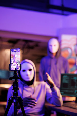 Smartphone screen with hackers recording threat video, asking victim for sensitive information and money. Criminals wearing anonymous masks threatening online and streaming fraud at night timeの写真素材