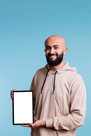 Smiling cheerful arab man holding digital tablet with white empty screen portrait. Carefree person showing portable device, displaying mock up for app ads and looking at cameraの写真素材