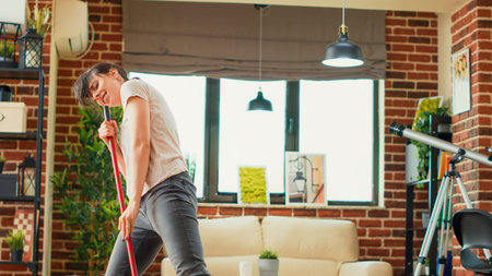 Young woman singing and dancing in living room, washing apartment floors with mop and washing...