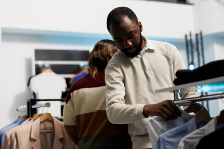 Shopper choosing formal wear outfit and searching for shirt size on rack in boutique. African...