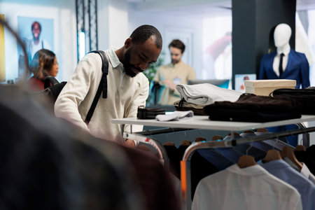 African american man browsing rack with casual clothes while shopping in mall fashion department. Boutique store customer examining menswear apparel while choosing trendy outfitの写真素材