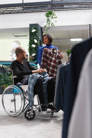 Clothing store asian woman customer showing plaid shirt on hanger to arab man with physical impairment. Mall boutique client in wheelchair getting assistance from seller while shopping for apparelの写真素材