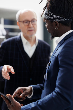 African american worker checking stylish shirt inventory on tablet computer for senior client in shopping centre. Elderly man buying formal clothes and fashionable merchandise in modern boutiqueの写真素材