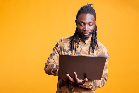 African american man holding laptop computer browsing webpages on internet, standing in studio over...