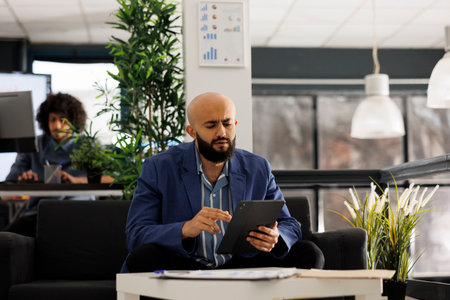 Arab executive manager checking financial data on tablet while working in business office. Startup company employee browsing internet on digital device and sitting on couch in coworking spaceの写真素材