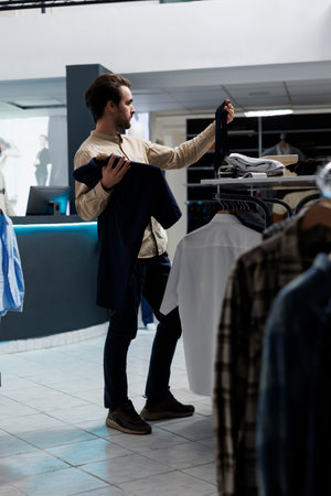 Clothing store customer selecting tie and shirt while shopping for formal outfit. Young man exploring fashion boutique while holding garment and accessory, checking styleの写真素材